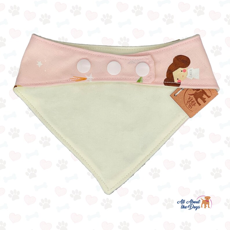 Peut inclure: Un bandana pour chien avec une section sup&eacute;rieure rose clair orn&eacute;e d'une illustration de dessin anim&eacute; et d'&eacute;toiles blanches. Le bandana a trois fermetures &agrave; pression blanches et une section inf&eacute;rieure cr&egrave;me. L'&eacute;tiquette indique "All About the Dogs".