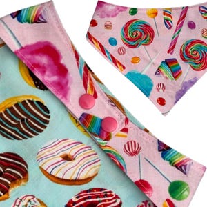 Pu&ograve; includere: Primo piano di due bavaglini per bambini a tema caramelle. Un bavaglino presenta uno sfondo azzurro con ciambelle, mentre l'altro ha uno sfondo rosa con lecca-lecca e bastoncini di zucchero. Entrambi i bavaglini hanno bottoni a pressione rosa.