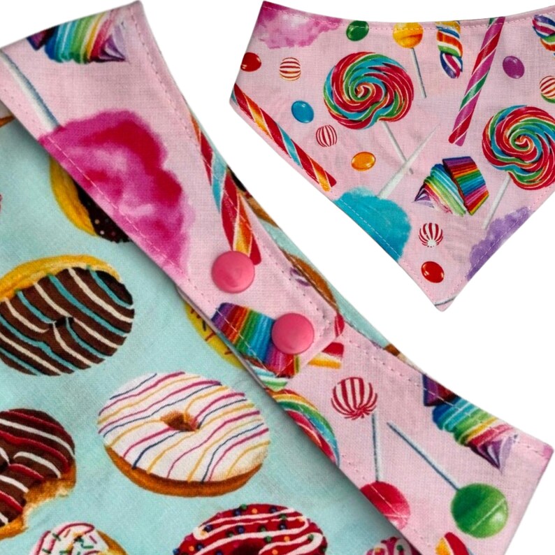 Pu&ograve; includere: Primo piano di due bavaglini per bambini a tema caramelle. Un bavaglino presenta uno sfondo azzurro con ciambelle, mentre l'altro ha uno sfondo rosa con lecca-lecca e bastoncini di zucchero. Entrambi i bavaglini hanno bottoni a pressione rosa.