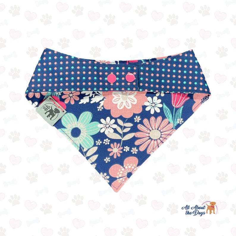 May include: A blue dog bandana with a floral pattern in pink, white, and teal. The top section features a navy blue polka dot design with pink buttons. A tag with a dog silhouette is attached.