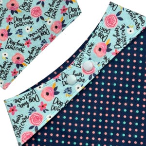 May include: Close-up of a dog bandana with a light blue floral print featuring the text "Dog hair, don't care." The bandana has a navy blue polka dot pattern on the reverse side and white snap closures.