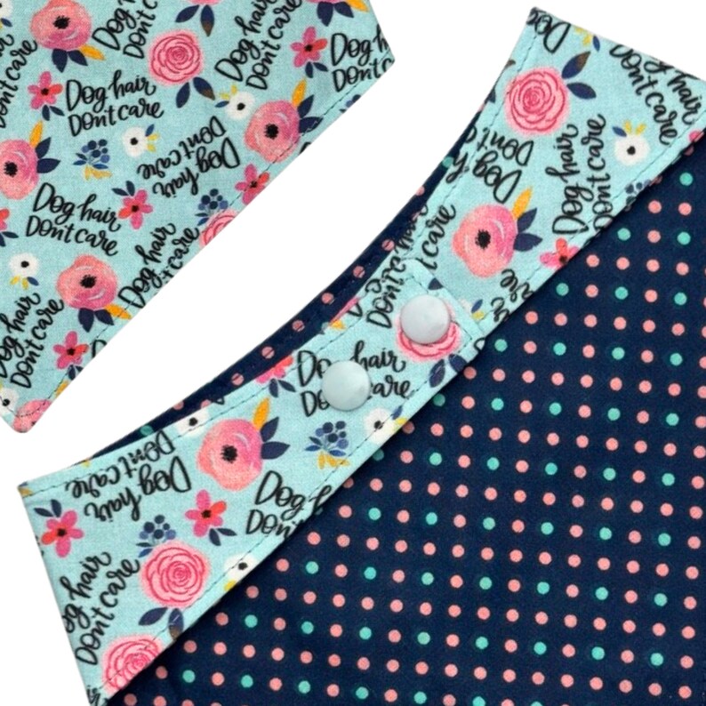 May include: Close-up of a dog bandana with a light blue floral print featuring the text "Dog hair, don't care." The bandana has a navy blue polka dot pattern on the reverse side and white snap closures.