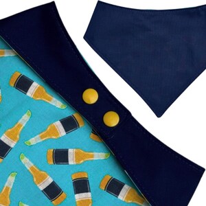 May include: A dog bandana with a blue and yellow beer bottle pattern. The bandana has a navy blue trim and two yellow snap closures. A separate solid navy blue bandana is also shown.