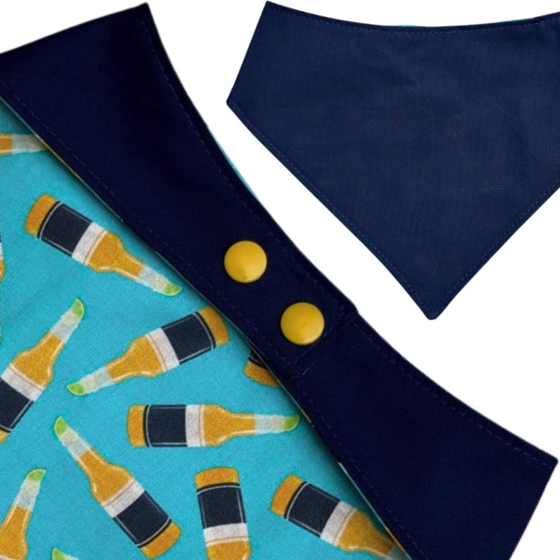 May include: A dog bandana with a blue and yellow beer bottle pattern. The bandana has a navy blue trim and two yellow snap closures. A separate solid navy blue bandana is also shown.