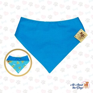 May include: A turquoise blue dog bandana with a white button and a pattern of green baby Yoda characters on the inside. The bandana is on a white background with a pattern of pink hearts and gray paw prints.