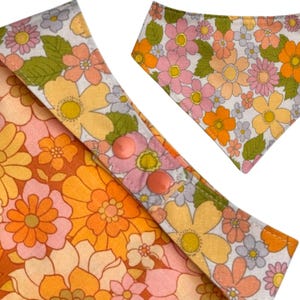 Puede incluir: Primer plano de dos art&iacute;culos de tela con estampado floral. Uno es un babero con cierre de bot&oacute;n a presi&oacute;n, con flores naranjas, amarillas, rosas y blancas. El otro es un babero triangular con el mismo estampado floral. La tela tiene una est&eacute;tica retro y vintage.