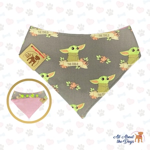 Puede incluir: Un bandana para perro gris y rosa con un patr&oacute;n repetido de un personaje alien&iacute;gena verde con el texto "The Child" en una pancarta. El bandana tiene una parte inferior rosa y una etiqueta marr&oacute;n con una silueta de perro.