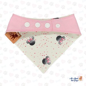 May include: Pink and white dog bandana with a Minnie Mouse design. The bandana has a pink top with three white snap closures. The bottom portion is white with a pattern of red dots and Minnie Mouse heads.