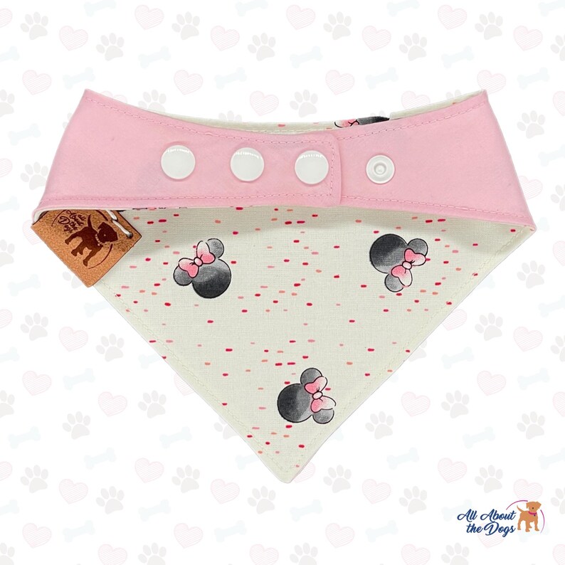 May include: Pink and white dog bandana with a Minnie Mouse design. The bandana has a pink top with three white snap closures. The bottom portion is white with a pattern of red dots and Minnie Mouse heads.