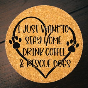 Puede incluir: Posavasos redondo de corcho con el texto negro "I JUST WANT TO STAY HOME DRINK COFFEE & RESCUE DOGS" dentro de un corazón con huellas de patas. El posavasos es de color marrón claro y está sobre una superficie de madera oscura.