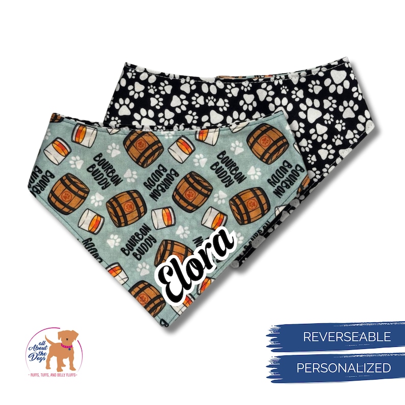 May include: Two reversible dog bandanas. One is light blue with bourbon barrels, paw prints, and the text "Bourbon Buddy". The other is black with white paw prints. The name "Elora" is on the light blue bandana.