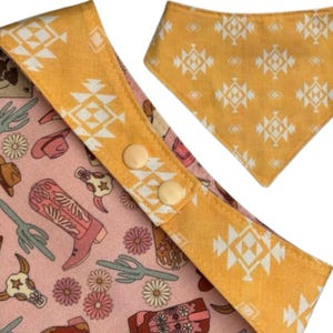 May include: A dog bandana and collar set with a pink and yellow southwestern design. The pink fabric features cowboy boots, cacti, and floral patterns. The yellow fabric has a white geometric design and the collar has two beige buttons.