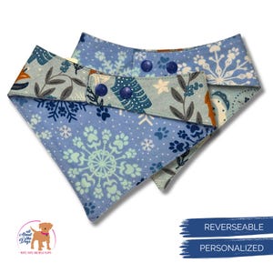 May include: Two reversible dog bandanas. One is light blue with a snowflake and paw print pattern. The other side is a light blue with a floral and tree design. The bandanas have blue snap closures. The text on the image says "REVERSIBLE" and "PERSONALIZED".