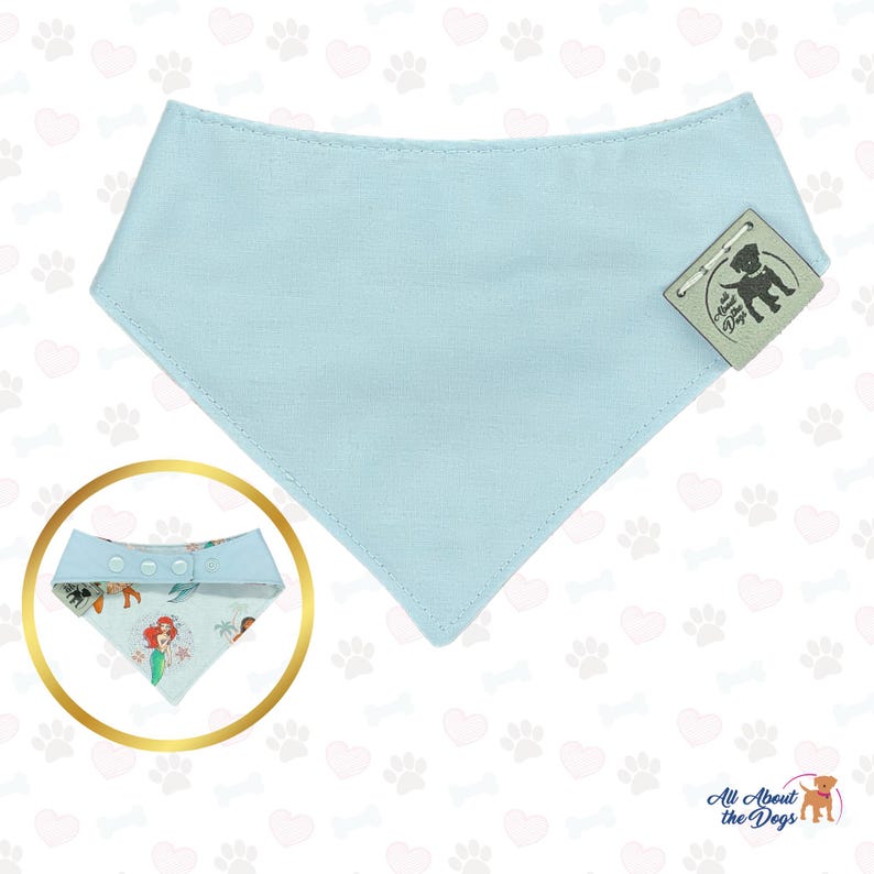 Peut inclure: Un bandana bleu clair pour chien, de forme triangulaire. Le bandana porte une &eacute;tiquette avec l'inscription "All About the Dogs". Un bandana plus petit avec une impression color&eacute;e d'une sir&egrave;ne et d'autres personnages est &eacute;galement visible.