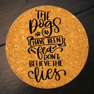 以下が含まれることがあります： 「THE DOGS HAVE BEEN FED DON'T BELIEVE THE LIES」の黒い文字が書かれた丸いコルクコースター。 コースターは明るい茶色で、表面はざらざらしています。 足跡のグラフィックが含まれています。