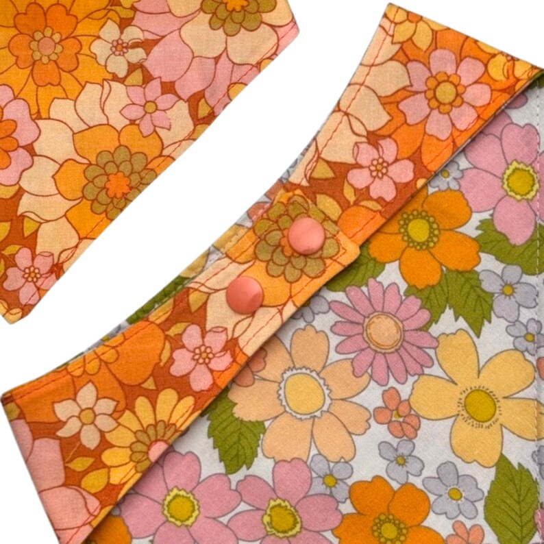 Puede incluir: Primer plano de una tela estampada con flores en tonos naranja, amarillo, rosa y verde. La tela presenta un dise&ntilde;o floral retro con dos botones rosas. La tela probablemente se utiliza para un babero o un art&iacute;culo similar.