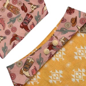 May include: Pink and yellow patterned dog bandana with a western theme. The pink side features cowboy boots, hats, and cacti. The yellow side has a geometric design. Two cream-colored buttons secure the bandana.