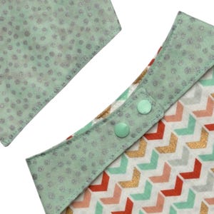 May include: Close-up of a baby bib with a mint green trim and two mint green snaps. The bib features a chevron pattern in red, gold, and mint green. The trim has a silver glitter dot pattern.