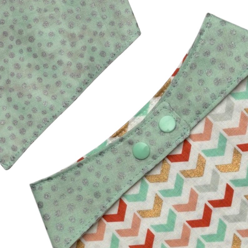 May include: Close-up of a baby bib with a mint green trim and two mint green snaps. The bib features a chevron pattern in red, gold, and mint green. The trim has a silver glitter dot pattern.