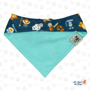 May include: A turquoise blue dog bandana with a dark blue border featuring a Star Wars themed print with characters like R2-D2, Chewbacca, and a Jawa. The bandana has two white snap buttons and a small brown tag with the text "All About the Dogs" in white.