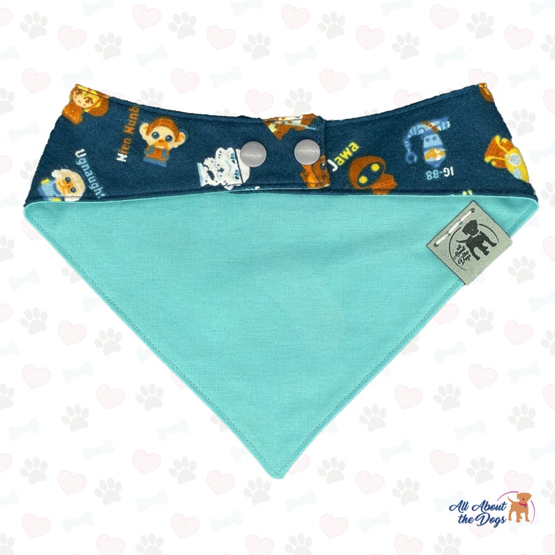 May include: A turquoise blue dog bandana with a dark blue border featuring a Star Wars themed print with characters like R2-D2, Chewbacca, and a Jawa. The bandana has two white snap buttons and a small brown tag with the text "All About the Dogs" in white.
