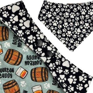 May include: Dog bandana featuring a black and white paw print design and a light blue bandana with bourbon barrel and glass graphics. The bandana has black snap closures.