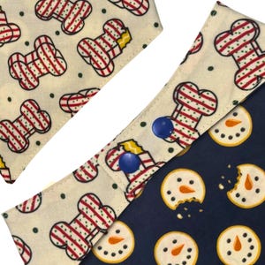 May include: Dog bandanas with festive patterns. One bandana features a bone design with red and white stripes on a cream background. Another has snowman cookies on a navy blue background. Both bandanas have blue snap closures.