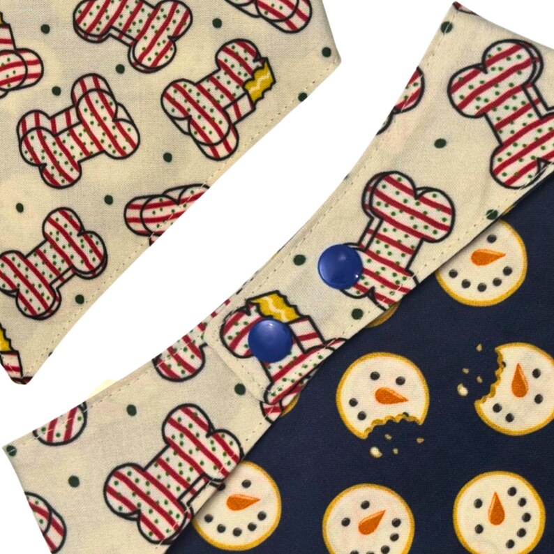 May include: Dog bandanas with festive patterns. One bandana features a bone design with red and white stripes on a cream background. Another has snowman cookies on a navy blue background. Both bandanas have blue snap closures.
