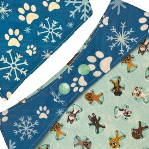 K&ouml;nnte beinhalten: Nahaufnahme eines Hunde-Bandanas mit blauem Hintergrund. Das Bandana zeigt wei&szlig;e Pfotenabdr&uuml;cke und Schneeflocken. Die R&uuml;ckseite ist hellblau mit Hundeillustrationen. Zwei hellgr&uuml;ne Kn&ouml;pfe sichern das Bandana.