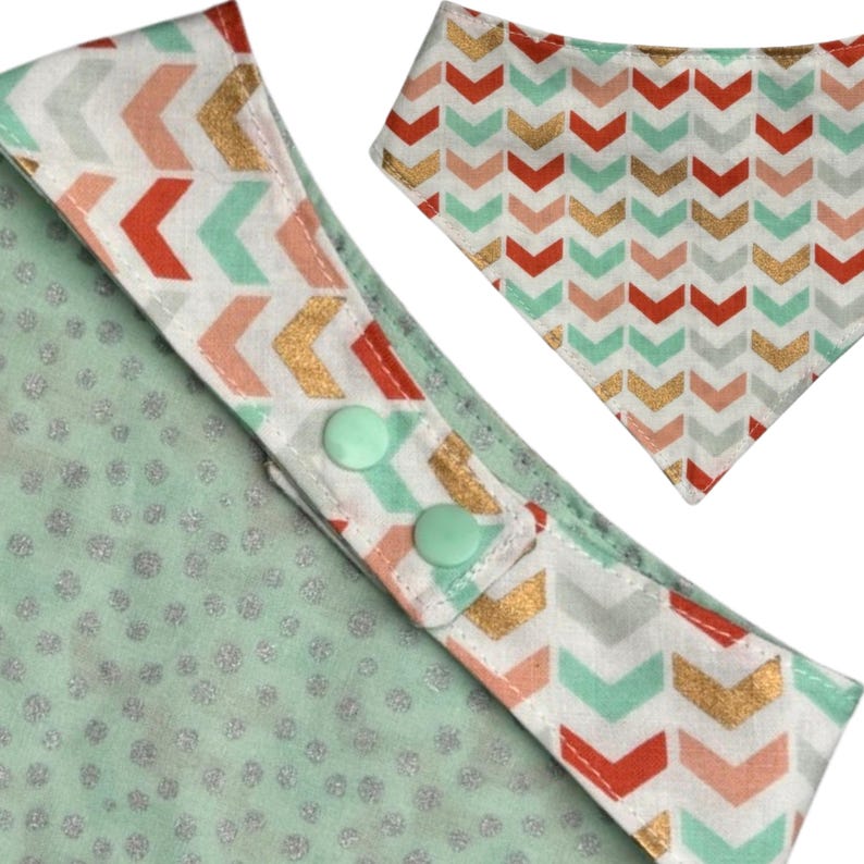 May include: A baby bib with a mint green background and a white border featuring a chevron pattern in red, gold, and mint green. The bib has two mint green snap closures. A matching bib is also visible.