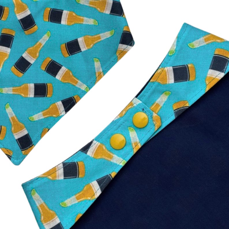 May include: A blue pet bandana and collar with a repeating pattern of beer bottles in yellow, black, and white. The collar has two yellow snap closures and a solid navy blue backing.