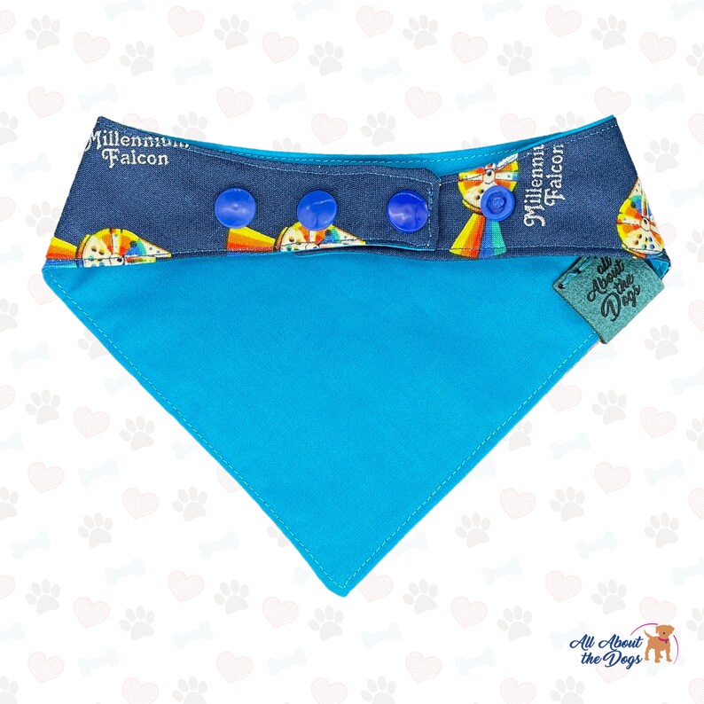 May include: A turquoise blue dog bandana with a navy blue and white patterned trim. The trim features a rainbow design and the text "Millennium Falcon". The bandana has three blue snap buttons and a green tag that says "All About the Dogs".