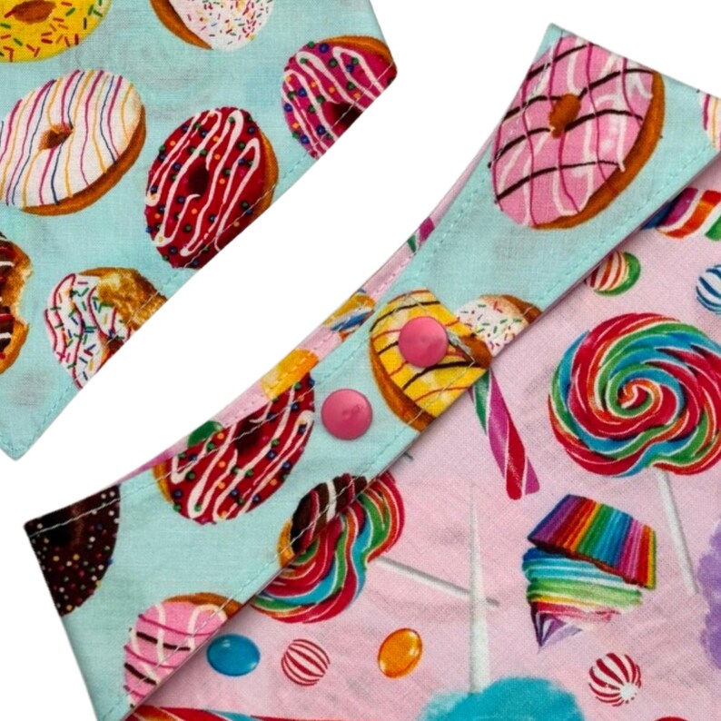Pu&ograve; includere: Primo piano di due pezzi di tessuto con un motivo colorato di ciambelle e lecca lecca. Un pezzo &egrave; azzurro con ciambelle, l'altro &egrave; rosa con lecca lecca. Bottoni rosa fissano i pezzi.