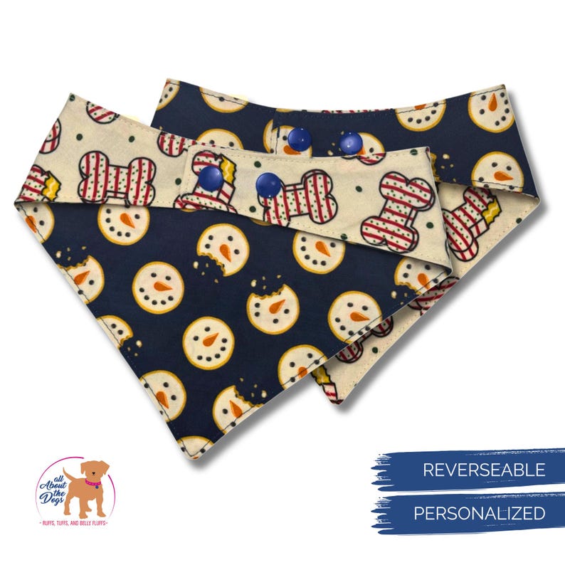 May include: Reversible dog bandana with a navy blue and cream pattern. One side features snowman cookies, the other side has a bone design. The bandana has blue snap closures. The text "REVERSIBLE" and "PERSONALIZED" are visible.