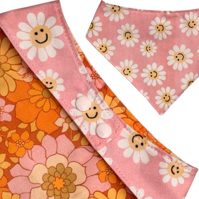 Puede incluir: Un conjunto de bandana y collar para perros con estampado floral rosa y naranja. El collar presenta margaritas blancas con caras sonrientes sobre un fondo rosa. La bandana tiene el mismo patr&oacute;n de margaritas. El collar tiene dos botones blancos.