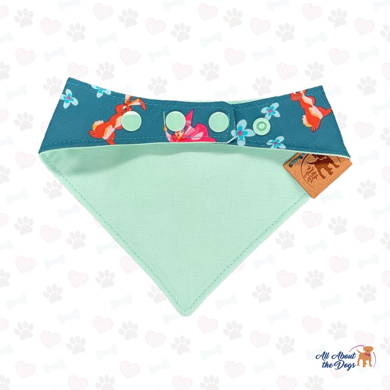May include: A teal dog bandana with a mint green underside. The top features a floral and rabbit pattern with snap closures. A small brown tag with the text "All About the Dogs" is attached.
