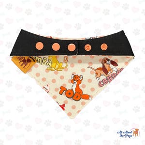 May include: A beige dog bandana with a black adjustable strap and pink snap buttons. The bandana features a white polka dot pattern and illustrations of characters from the Disney movie The Fox and the Hound, including Tod, Copper, and  a lion named  "Bumba".