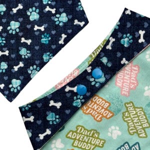 Puede incluir: Dos pa&ntilde;uelos para perros. Uno presenta un estampado de huellas de patas y huesos azul marino, el otro tiene un fondo azul claro con el texto "Dad's Adventure Buddy". Uno tiene cierres de bot&oacute;n a presi&oacute;n azules.