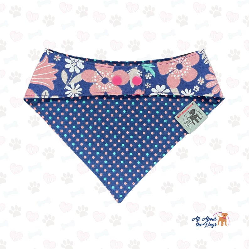 May include: A navy blue dog bandana with a floral pattern and pink polka dots. The top section features pink and white flowers on a navy background, secured with pink buttons. The bottom is navy with pink and blue polka dots. A small tag is attached.