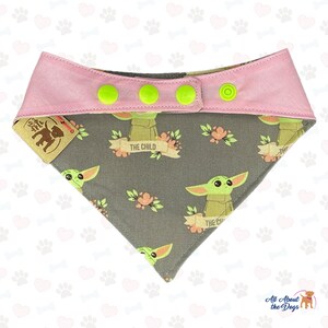 Puede incluir: Un bandana para perro gris y rosa con un patr&oacute;n repetido de un personaje alien&iacute;gena verde con el texto "The Child" en una pancarta. El bandana tiene cuatro botones a presi&oacute;n verdes.