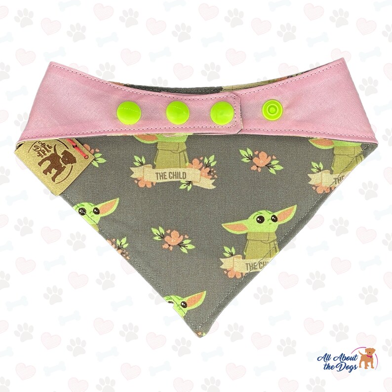 Puede incluir: Un bandana para perro gris y rosa con un patr&oacute;n repetido de un personaje alien&iacute;gena verde con el texto "The Child" en una pancarta. El bandana tiene cuatro botones a presi&oacute;n verdes.