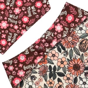 May include: Close-up of a dog bandana with a floral pattern and the words "Wild Thing". The bandana has a brown border with pink flowers and paw prints. The interior has a cream background with pink and orange flowers.