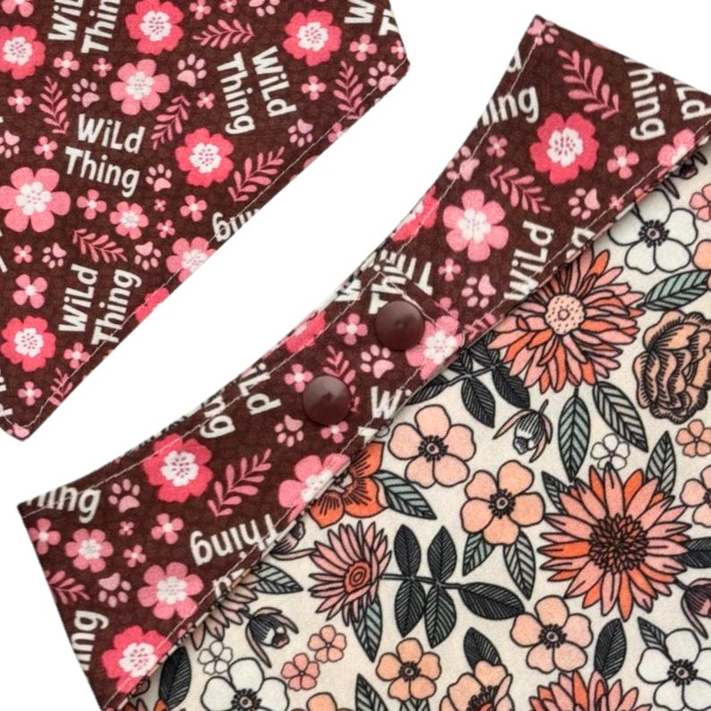 May include: Close-up of a dog bandana with a floral pattern and the words "Wild Thing". The bandana has a brown border with pink flowers and paw prints. The interior has a cream background with pink and orange flowers.