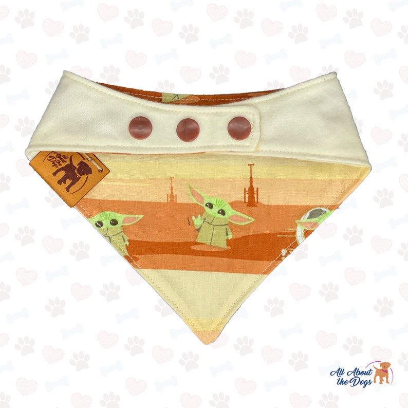 May include: A dog bandana featuring a Star Wars theme with a Grogu character design. The bandana has a cream-colored top with three brown buttons and a patterned bottom with Grogu illustrations in a desert landscape. A small tag with a dog logo is attached.