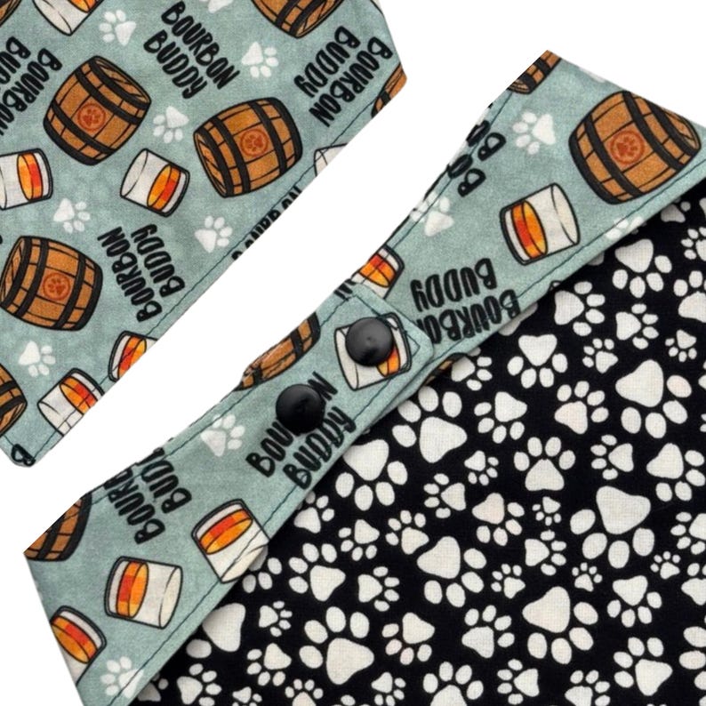 May include: A dog bandana with a light blue background featuring brown bourbon barrels, whiskey glasses, and the text "BOURBON BUDDY." The reverse side is black with white paw prints. The bandana has two black snap closures.