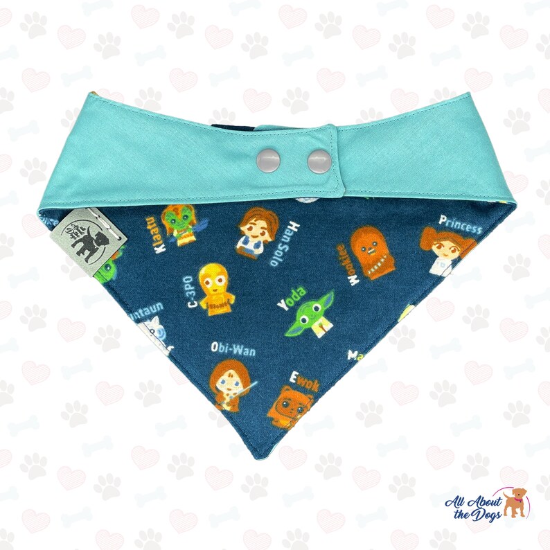 May include: A blue and teal dog bandana with a Star Wars theme. The bandana features a variety of Star Wars characters, including Yoda, Chewbacca, Princess Leia, and C-3PO. The bandana has a snap closure.