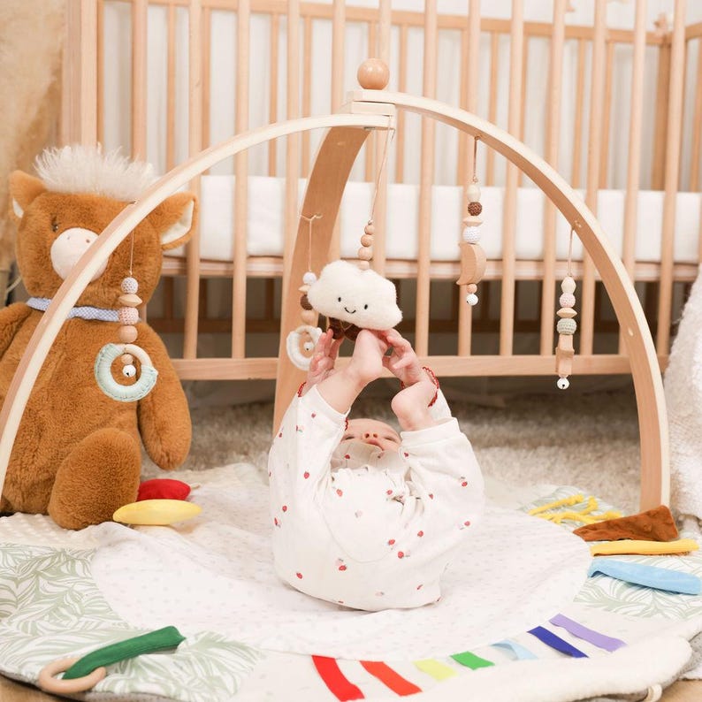 Pode incluir: Um gin&aacute;sio de madeira para beb&eacute;s com brinquedos suspensos, uma nuvem de pel&uacute;cia e um tapete de brincar macio com detalhes coloridos. Um grande urso de pel&uacute;cia e um ber&ccedil;o de madeira est&atilde;o no fundo. O beb&eacute; est&aacute; a usar um body branco com bolinhas vermelhas.