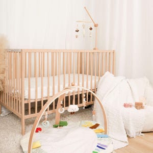 Pode incluir: Um ber&ccedil;o de madeira clara com um colch&atilde;o branco, um gin&aacute;sio de madeira para beb&eacute;s com brinquedos suspensos e um m&oacute;bile de madeira com formas suspensas. Um cobertor e uma almofada brancos est&atilde;o no ch&atilde;o. O quarto tem uma paleta de cores neutras.