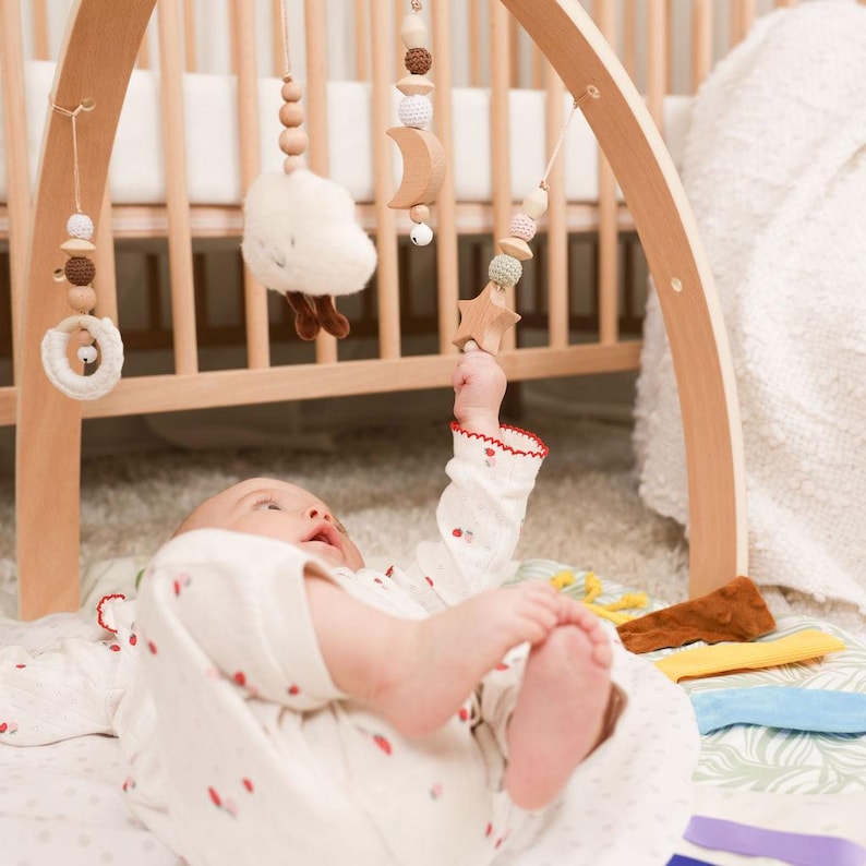 Pode incluir: Um gin&aacute;sio de madeira para beb&eacute;s com brinquedos suspensos, incluindo uma nuvem, uma lua e uma estrela. Os brinquedos est&atilde;o suspensos sobre um beb&eacute; deitado num tapete de brincar com padr&otilde;es. O beb&eacute; est&aacute; a usar um body branco com detalhes vermelhos.