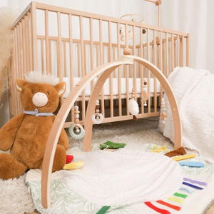 Pode incluir: Um gin&aacute;sio de madeira para beb&eacute;s com brinquedos suspensos, um tapete de brincar macio e um urso de peluche. O gin&aacute;sio tem uma estrutura arqueada com brinquedos de madeira e tecido pendurados. Um ber&ccedil;o de madeira est&aacute; no fundo.